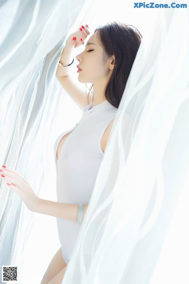 A woman in a white bodysuit leaning against a white curtain.
